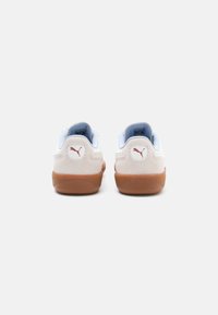 Zapatilla blanca y azul claro con talón de ante, suela de goma y logo rojo en la parte trasera. Textura suave y diseño redondeado.