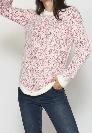 Femme portant un pull duveteux blanc et rose tacheté avec un jean bleu foncé, une main dans la poche, debout devant un fond uni.