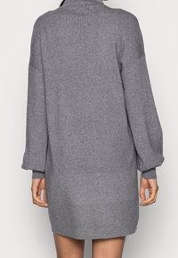 Personne portant une robe-pull en maille grise à manches longues et longueur genou, vue de dos sur un fond uni.