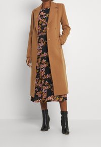 Lång, kamelfärgad kappa i en ullblandning med dubbelknäppt design, lagd över en svart blommig klänning, tillsammans med svarta ankelstövlar.