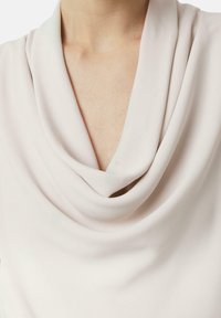 Gros plan sur le col d'un chemisier beige drapé, visible sur le haut de la poitrine et le cou d'une personne.