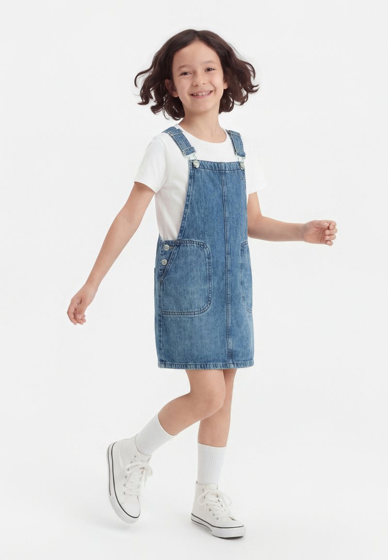 Νεαρή κοπέλα που χαμογελά, φοράει λευκό μπλουζάκι, φόρεμα με τιράντες από τζιν, λευκές κάλτσες και λευκά αθλητικά παπούτσια, στέκεται μπροστά σε απλό λευκό φόντο.