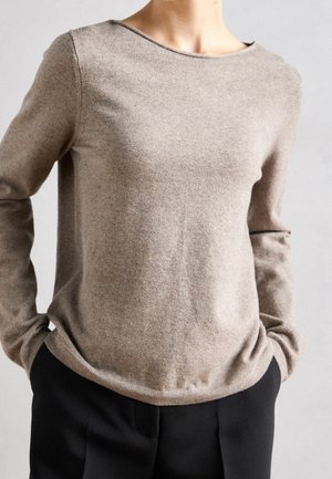 Person iført en beige langærmet rundhalset sweater, let stoppet ned i sorte bukser med hænderne i lommerne, neutralt baggrund.