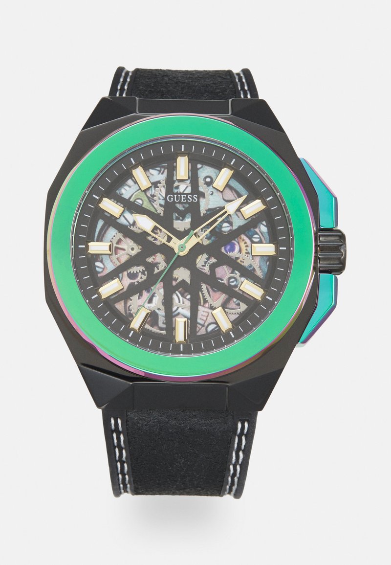 Guess Uhr mit achteckigem schwarzem Gehäuse, grüner Lünette, transparentem Ziffernblatt, das die Uhrwerke zeigt, goldenen Stundenmarkierungen und schwarzem Lederarmband mit weißen Nähten.