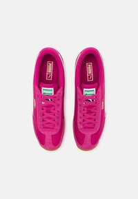 Kirkas vaaleanpunainen tennari, jossa on mokkanahkaa ja kangasta yläosa, varustettuna vastaavilla nauhoilla, valkoisilla yksityiskohdilla ja kumikauttaalla. PUMA-logo kielekkeessä.