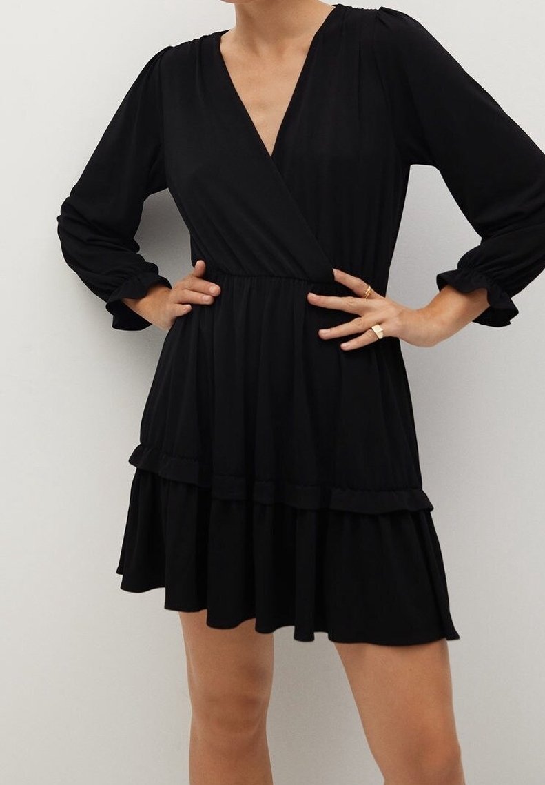 Personne portant une robe noire à manches longues, col en V, au-dessus du genou avec un ourlet à volants, les mains sur les hanches, debout contre un mur clair uni.