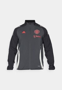 Chaqueta deportiva negra con el logotipo rojo de Manchester United y la marca Tezos. Presenta paneles laterales blancos y tres franjas blancas en los hombros.