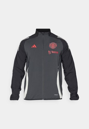 Chaqueta deportiva negra con el logotipo rojo de Manchester United y la marca Tezos. Presenta paneles laterales blancos y tres franjas blancas en los hombros.
