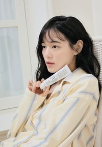 En hvit kremtube merket "147 Barrier Cream" holdt mot ansiktet til en person som har på seg en overdimensjonert skjorte med gul- og blåstriper.