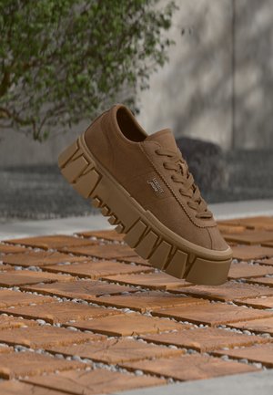 Baskets en daim marron avec une semelle épaisse et robuste et des lacets flottant au-dessus d'une surface extérieure en bois et en carreaux de pierre.