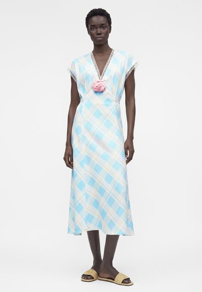 Γυναίκα που φοράει φόρεμα μεσαίου μήκους σε ανοιχτό μπλε και λευκό καρό με ροζ υφασμάτινο λουλούδι στο ντεκολτέ και μπεζ σανδάλια.