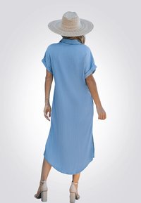 Vestido de camisa azul claro, hasta la rodilla, con mangas cortas dobladas, un cuello y aberturas laterales. Lleva un sombrero de paja de ala ancha y tacones cuadrados beiges.