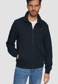 Sweatshirt zippé bleu marine en tissu doux avec un col haut, doté de poches latérales et d'un simple logo sur le côté gauche de la poitrine.