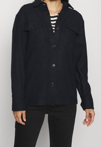 Navy napapaita, jossa on kaksi etutaskua, muotoiltu kaulus, pitkät hihat ja pehmeä, teksturoitu kangas. Yhdistetty mustiin housuihin.