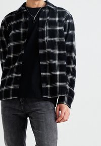Svart och vit rutig flanellskjorta med knapp-up design, med en bröstficka, bärs över en svart t-shirt, tillsammans med mörka jeans.