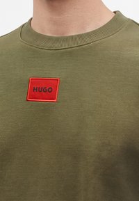 Κοντινή λήψη ενός πράσινου μπλουζιού με στρογγυλή λαιμόκοψη και ένα κόκκινο ορθογώνιο ταμπελάκι που εμφανίζει το όνομα της μάρκας "HUGO" με μαύρα γράμματα.