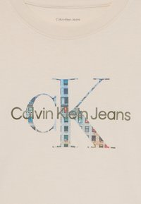 Krémový bavlnený tričko s veľkým, štýlizovaným logom "CK" s vzorom budov a textom "Calvin Klein Jeans" v tmavosivej farbe.