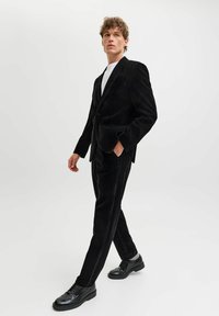 Juodas kordurovinis kostiumas, sudarytas iš prigludusio švarko su iškirpto atlapu ir tiesių kojų kelnių. Dėvimas su juodais raišteliais užsegamais batais ir balta marška.