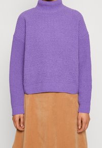 Violetti neulottu neulepaita korkealla kauluksella, resorikuvioisella pinnalla ja väljällä istuvuudella, yhdistetty ruskeaan corduroy-kankaiseen hameeseen.