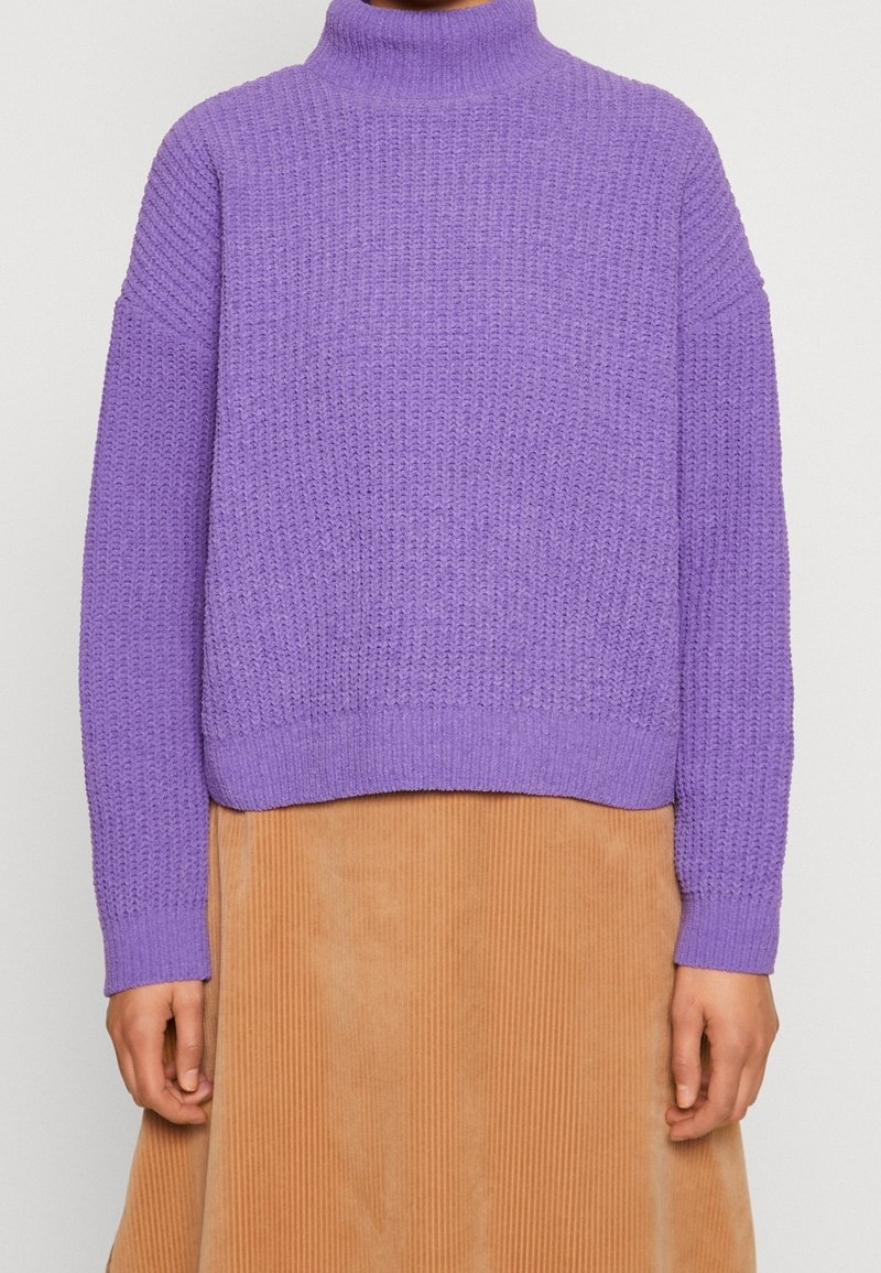 Violetti neulottu neulepaita korkealla kauluksella, resorikuvioisella pinnalla ja väljällä istuvuudella, yhdistetty ruskeaan corduroy-kankaiseen hameeseen.