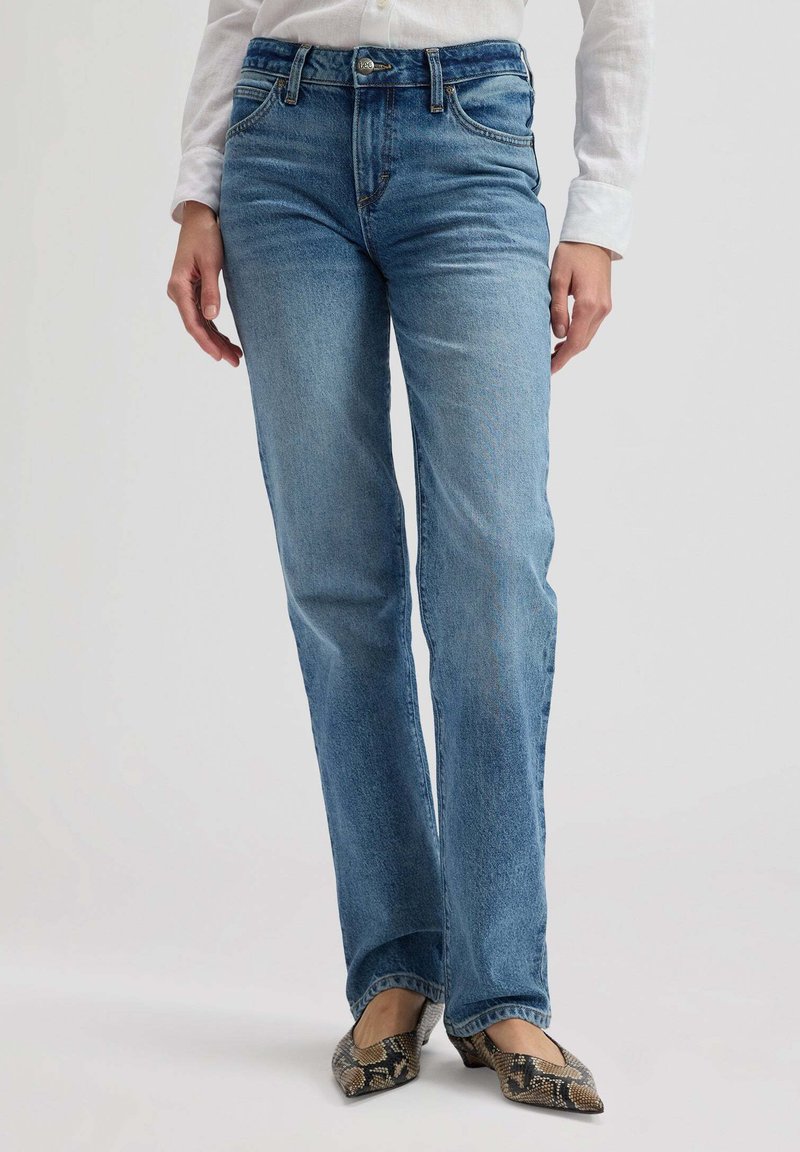 Άτομο που φορά ευθεία μπλε τζιν παντελόνια, λευκό πουκάμισο με μακριά μανίκια και μυτερά παπούτσια με σχέδιο φιδίσιας επιδερμίδας, στέκεται σε ανοιχτό φόντο.