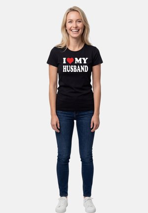 Lächelnde Frau, stehend, trägt ein schwarzes T-Shirt mit weißem Text und rotem Herzgrafik mit der Aufschrift "Ich liebe meinen Ehemann", blaue Jeans und weiße Turnschuhe.