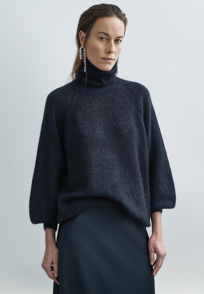 Dunkel marineblauer, oversized Rollkragenpullover mit flauschiger Textur, ausgestellten Schultern und dreiviertel Ärmeln. Das Model trägt einen eleganten schwarzen Rock.