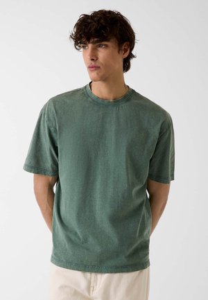 Młody mężczyzna z kręconymi włosami, ubrany w wyblakły zielony t-shirt z krótkim rękawem i jasne spodnie, patrzący w bok na tle jednolitej ściany.