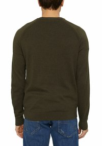 Homme portant un pull vert foncé à manches longues et un jean bleu, vu de dos sur un fond blanc uni.