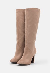 Par de botas de ante beige hasta la rodilla con puntas afiladas y tacones de bloque sobre un fondo blanco.
