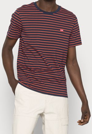 Homme portant un t-shirt bleu marine à fines rayures rouges horizontales et un pantalon beige clair, une main dans la poche.