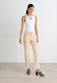 Top bianco senza maniche con logo, jeans beige a gamba dritta e sandali neri con tacco a blocco caratterizzati da un audace motivo di testo.