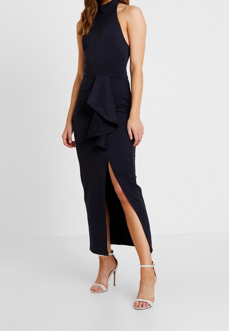 Femme portant une robe noire sans manches avec une fente haute et un détail à volants, associée à des sandales blanches à talons hauts.