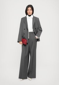 Blazer grigio doppiopetto abbinato a una camicia bianca e pantaloni grigi a vita alta. Tiene in mano una piccola borsa rossa e indossa décolleté bianche a punta.