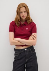 Rotes, geripptes Poloshirt mit kurzärmeligen, drei-Knopf-Leiste und gesticktem weißen Logo; kombiniert mit hoch geschnittenen, dunklen Jeans.