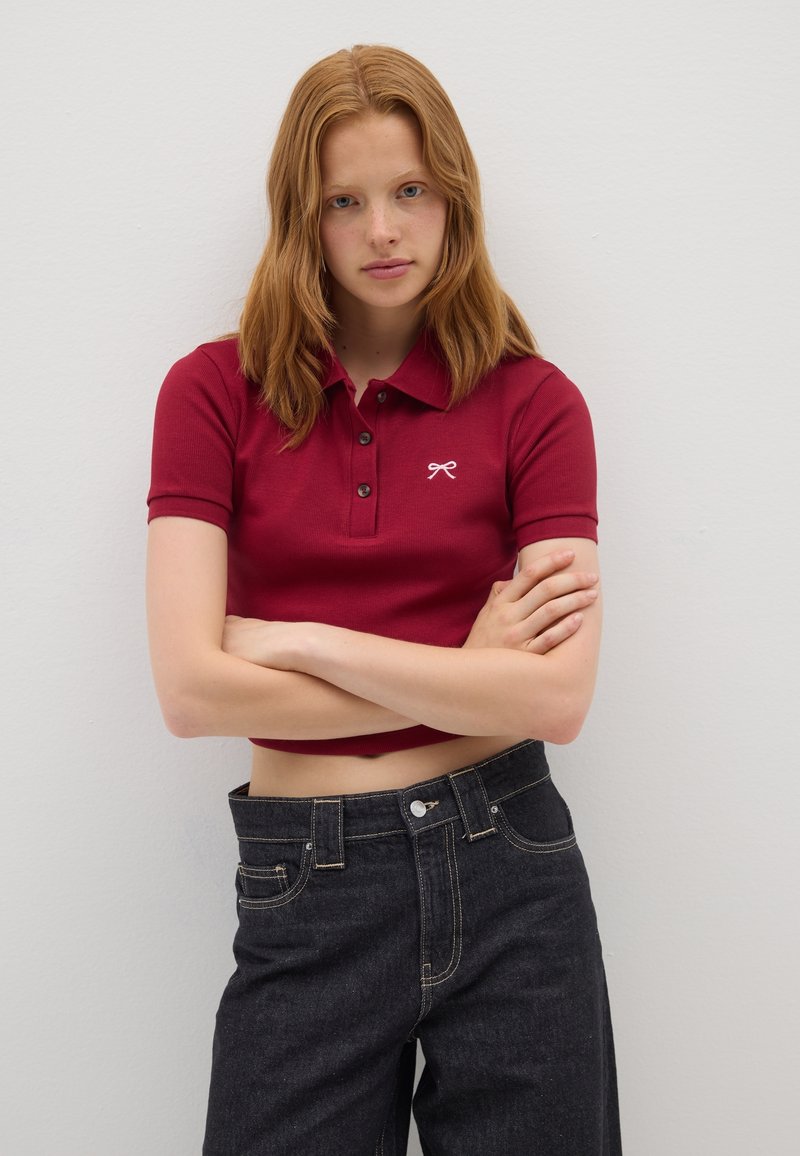 Rotes, geripptes Poloshirt mit kurzärmeligen, drei-Knopf-Leiste und gesticktem weißen Logo; kombiniert mit hoch geschnittenen, dunklen Jeans.