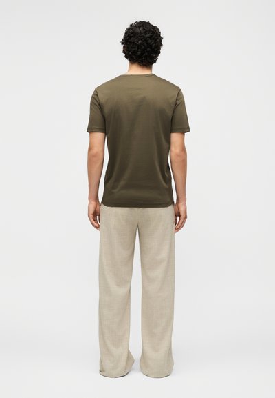 Άνδρας με σγουρά μαλλιά, που φοράει ελαιοπράσινο κοντομάνικο πουκάμισο και μπεζ παντελόνι φαρδιάς γραμμής, στέκεται γυρισμένος με την πλάτη σε ένα μονόχρωμο φόντο.