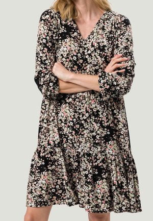 Vrouw draagt een zwarte jurk met lange mouwen met een kleine witte en roze bloemenprint, armen gekruist, staand tegen een effen lichte achtergrond.