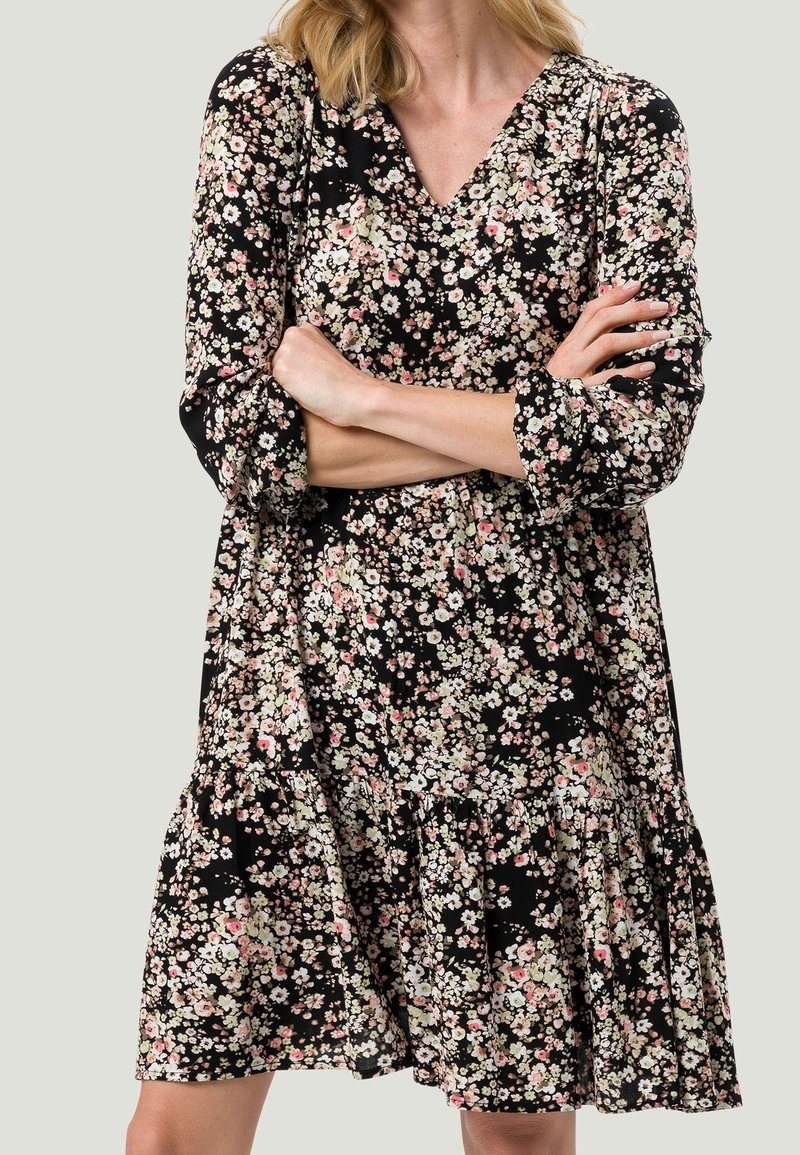Femme portant une robe noire à manches longues avec un petit imprimé floral blanc et rose, bras croisés, debout devant un fond clair uni.