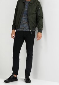 Mörkgrön bomberjacka med ett blixtlåsförsett ficka, kombinerad med en strukturerad blå tröja, svarta smala byxor och svarta sneakers.