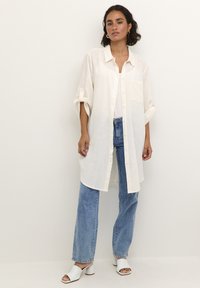 Chemise longue de couleur beige clair avec un devant à boutons, des manches retroussées et une poche à l'avant, associée à un jean bleu et des mules blanches à bout ouvert.