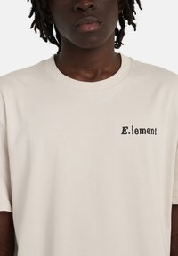 Μπεζ βαμβακερό T-shirt με άνετη εφαρμογή, κοντά μανίκια και τυπωμένο μαύρο λογότυπο με την επιγραφή "E.lement" στην αριστερή πλευρά του στήθους.