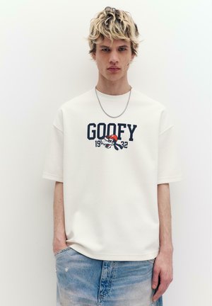 Giovane uomo con capelli biondi ricci indossa una maglietta bianca oversize con la scritta "Goofy 1932" e una grafica di cartone animato, abbinata a jeans azzurri.
