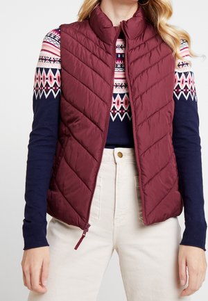 Femme portant une veste matelassée bordeaux sur un pull à motifs bleu marine et rose et un pantalon taille haute couleur écru, debout les bras le long du corps.