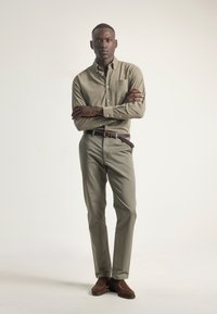 Hombre de pie con los brazos cruzados, vistiendo una camisa verde oliva de botones, pantalones a juego, un cinturón trenzado marrón y zapatos de ante marrones.