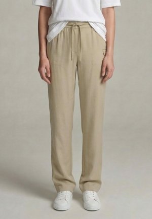 Person wearing beige drawstring pants, white T-shirt, and white sneakers standing against a plain light background.