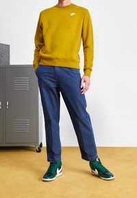 Gul bomullströja med en liten vit logotyp, ihop med blå denimbyxor och gröna sneakers med vita detaljer. Strukturerad yta.