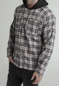 Camicia a quadri beige e navy, con bottoni frontali, due tasche sul petto e un cappuccio nero. Realizzata in un tessuto morbido e testurizzato.