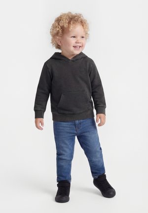 Usmievajúce sa batoľa s kučeravými blond vlasmi, oblečené v čiernej mikine s kapucňou, modrých džínsoch a čiernych teniskách, stojace na bielom pozadí.