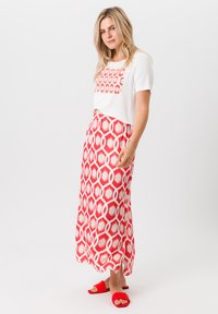 Witte T-shirt met een rood geometrisch patroon gecombineerd met een lange rode en witte gestreepte rok, aangevuld met rode platte sandalen.